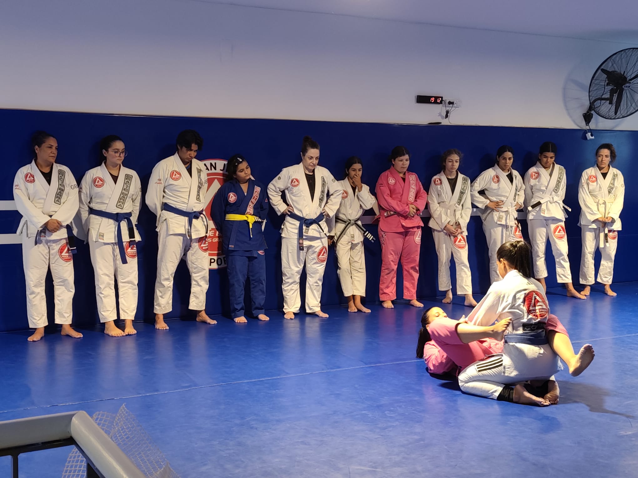 Turma feminina da Gracie Barra Braga alinhada junto à parede, com duas alunas a treinar uma técnica no tatame