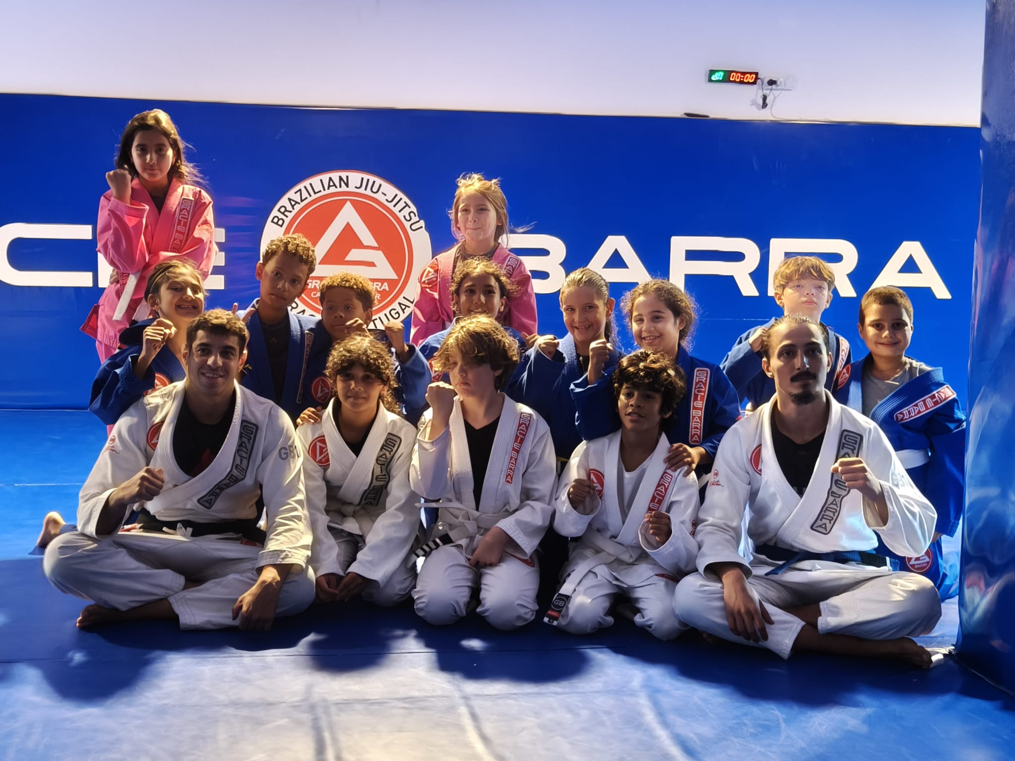Grupo de crianças da Gracie Barra Braga sentadas de kimono, com o punho fechado, à frente da parede Gracie Barra