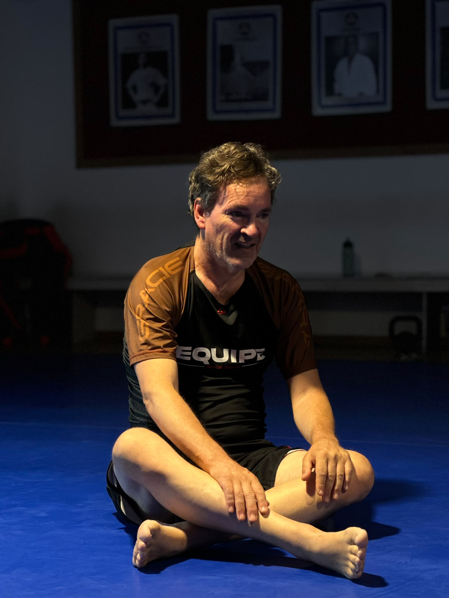 Instrutor da Gracie Barra Braga sentado no tatame, com fotografias emolduradas de mestres de Jiu-Jitsu na parede